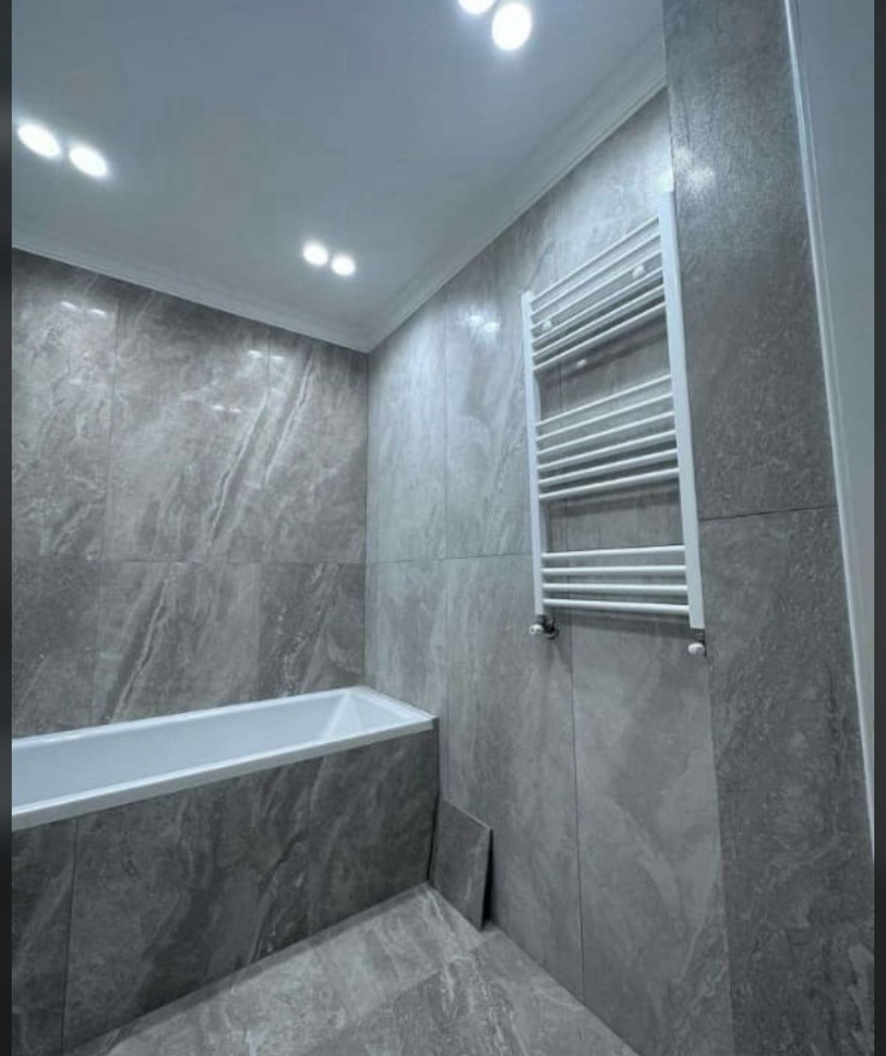 Wykończona łazienka w szarościach: marmurowe płytki na ścianach i podłodze, biała wanna i grzejnik drabinkowy. Nowoczesne oświetlenie sufitowe.