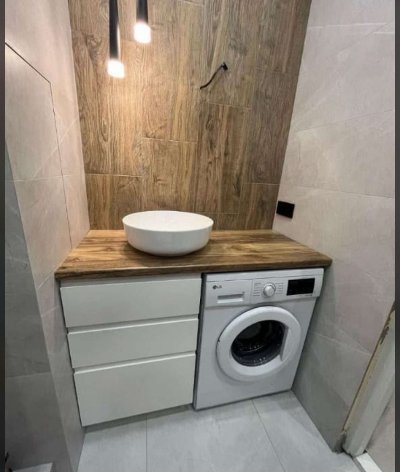 Nowoczesna łazienka z drewnianą ścianą, blatem i białą umywalką nablatową. Obok pralka i białe szafki z szufladami. Minimalistyczne oświetlenie.
