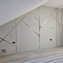 Usługi remontowo-budowlane - Ściana w pokoju na poddaszu z dekoracyjnymi, złotymi listwami ułożonymi w geometryczny wzór, widoczne puszki elektryczne bez osprzętu i podłoga z paneli imitujących drewno.