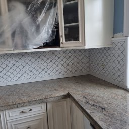 Usługi remontowo-budowlane - Kuchnia w trakcie odświeżania: białe szafki z kremowym blatem, mozaika na ścianie, zabezpieczona folią szafka wisząca, ślady szpachli przy ścianie.