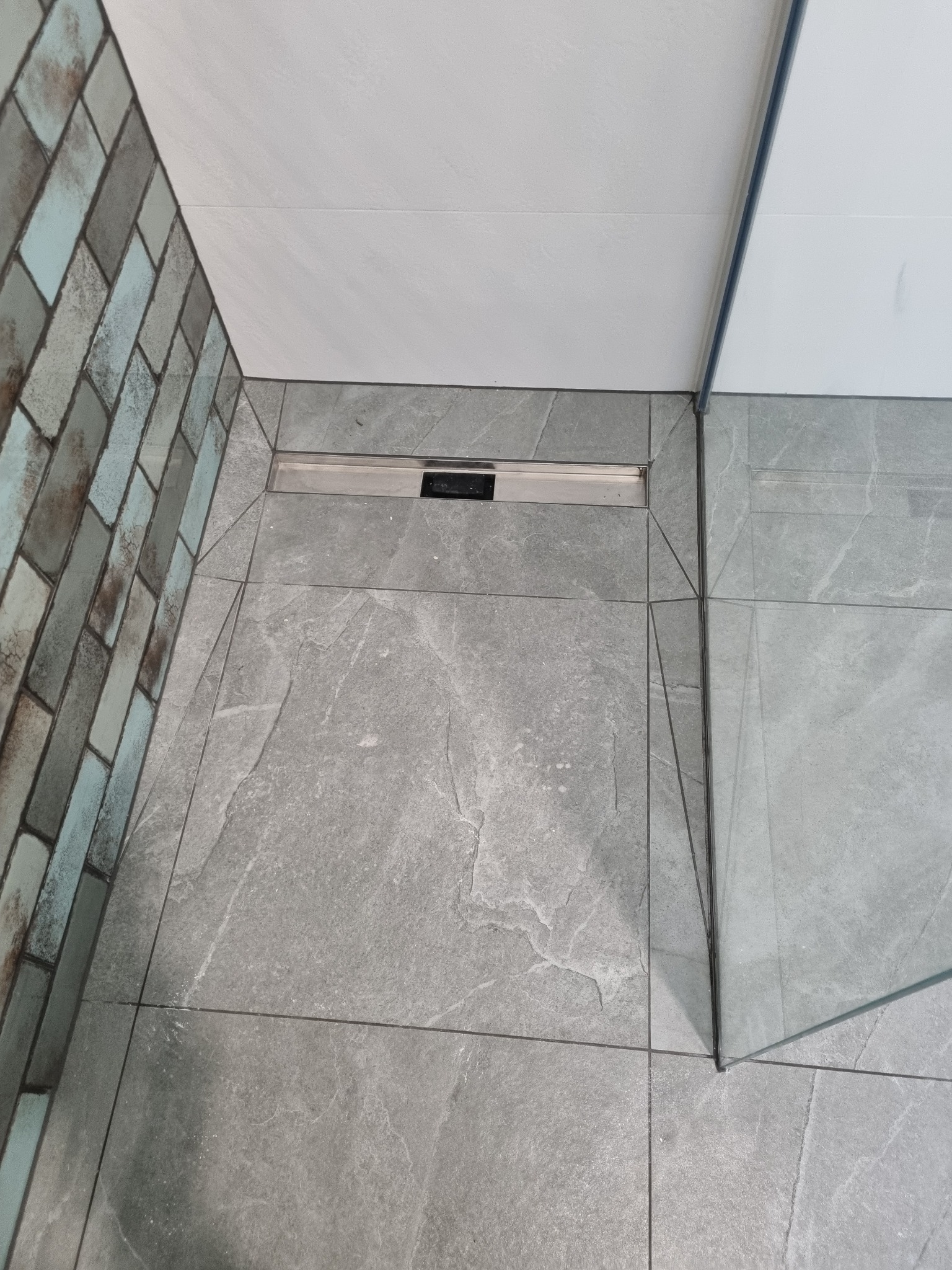 Nowoczesna łazienka: szare płytki podłogowe z imitacją kamienia, chromowany odpływ liniowy, ściana z dekoracyjnej cegły w odcieniach szarości i błękitu oraz fragment szklanej kabiny prysznicowej.