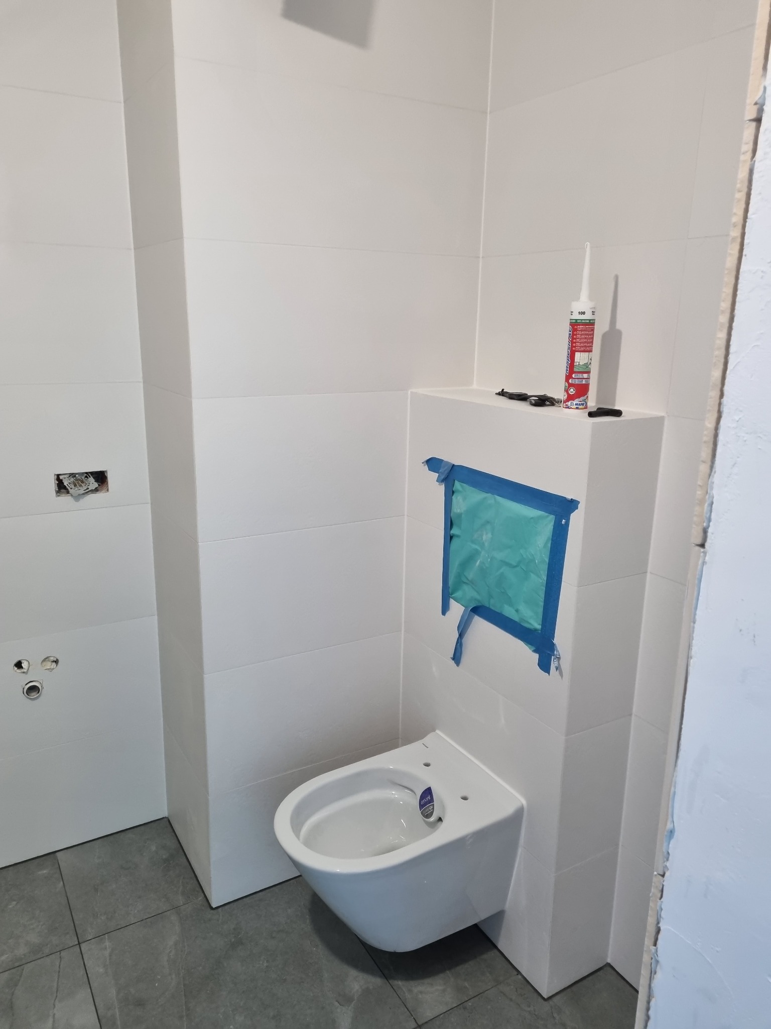 Biała, podwieszana toaleta w trakcie montażu w łazience z białymi płytkami, zabezpieczona niebieską folią i taśmą malarską, widoczna tuba silikonu i narzędzia na obudowie stelaża, szare płytki...