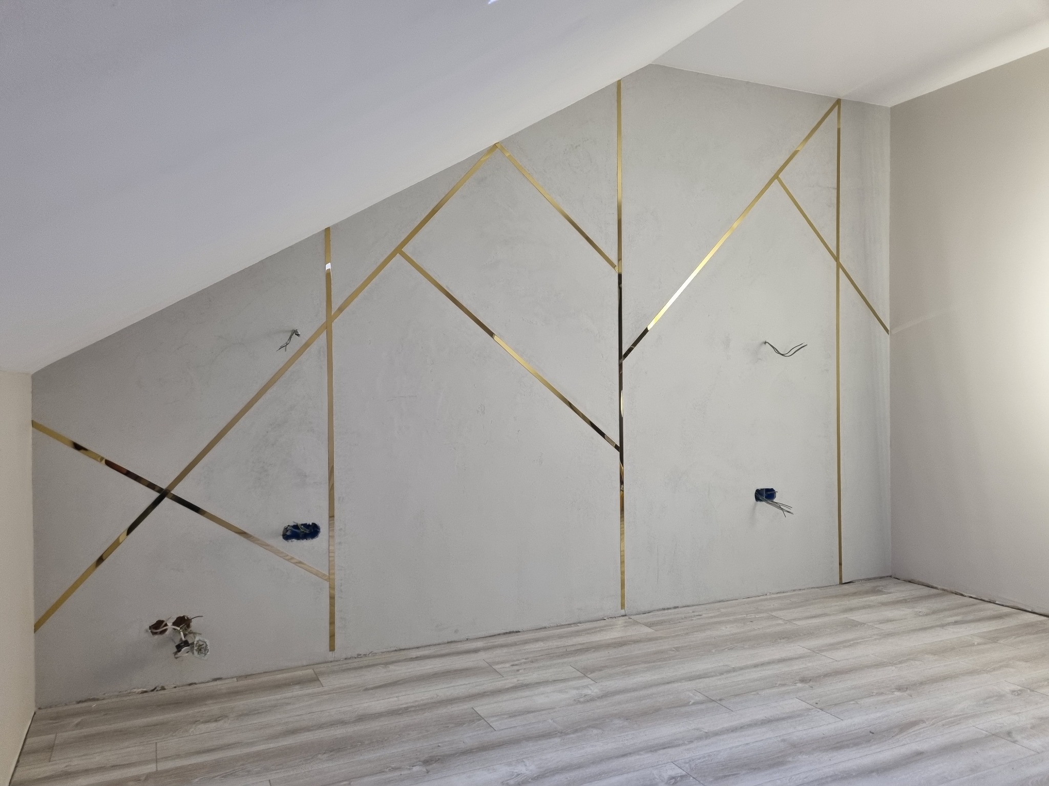 Ściana w pokoju na poddaszu z dekoracyjnymi, złotymi listwami ułożonymi w geometryczny wzór, widoczne puszki elektryczne bez osprzętu i podłoga z paneli imitujących drewno.