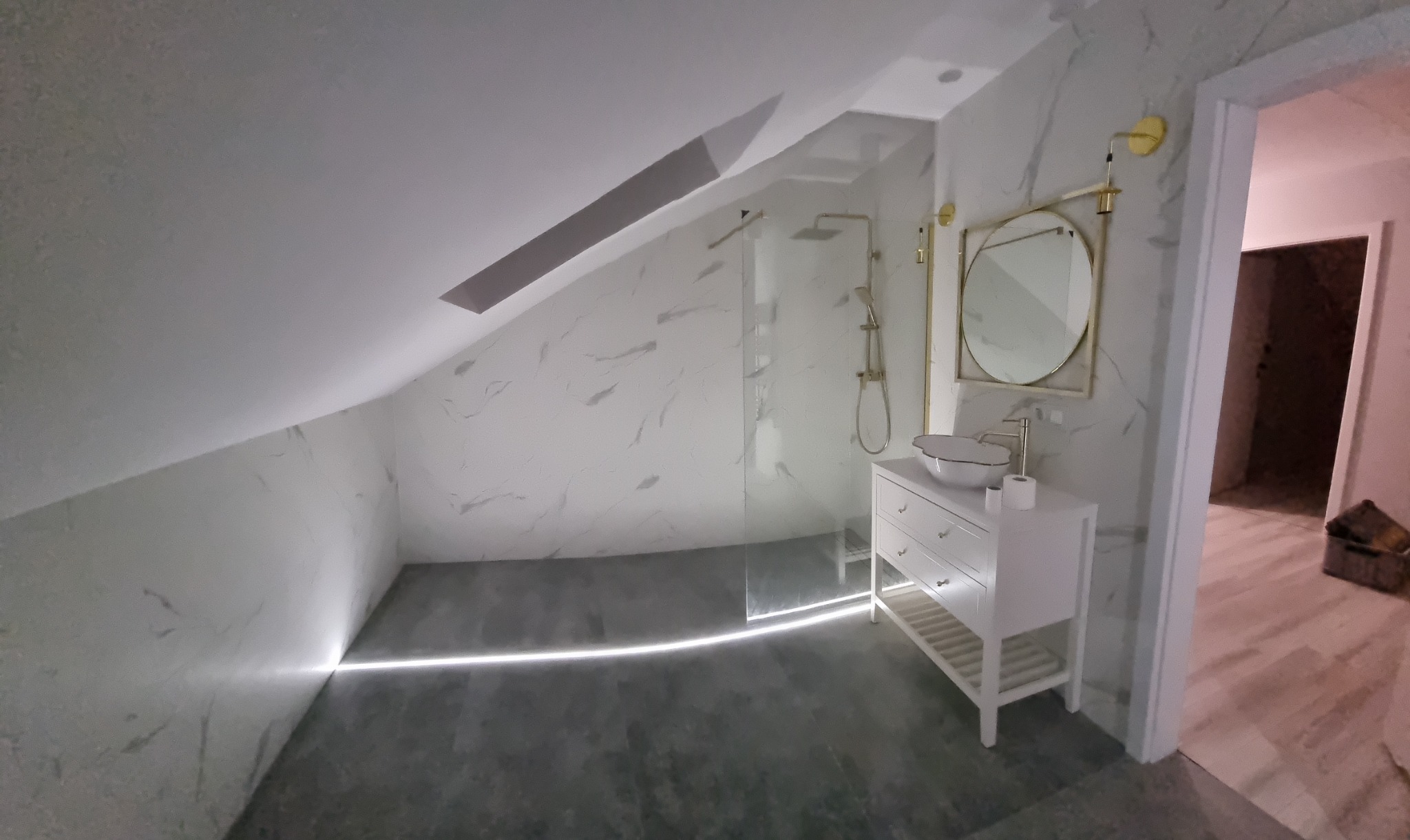 Nowoczesna łazienka na poddaszu ze skośnym sufitem, marmurowymi ścianami, kabiną prysznicową typu walk-in z złotą armaturą, białą szafką z umywalką nablatową i okrągłym lustrem ze złotą ramą...