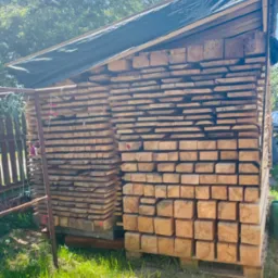 Stos tarcicy budowlanej i kantówki ułożony na paletach i przykryty plandeką, zabezpieczony metalową konstrukcją na zewnątrz.