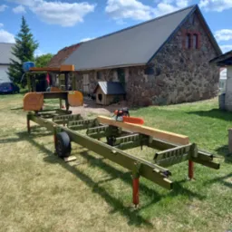 Mobilny tartak z piłą łańcuchową na belce, w tle budynek z kamienia i drewniana buda dla psa.