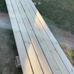 Stos świeżo obrobionych desek ułożonych na drewnianych podkładach na trawie, widoczne słoje drewna i delikatna poświata słoneczna.