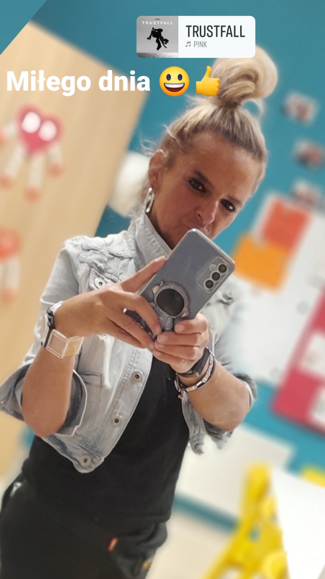 Osoba robi selfie telefonem z naklejką informującą o słuchanej muzyce, widoczne akcesoria i fragment wnętrza z dekoracjami.