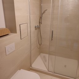 Huliailo Dmytro - Jasna łazienka po remoncie z kabiną prysznicową z przesuwnymi drzwiami, podwieszaną toaletą i ukrytym zaworem. Na ścianie widoczny właz rewizyjny.