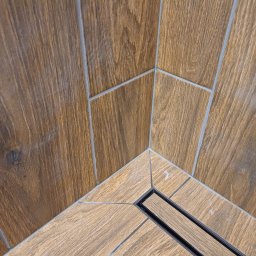 Huliailo Dmytro - Fragment wykończonej kabiny prysznicowej z odpływem liniowym, wyłożonej płytkami imitującymi drewno w dwóch odcieniach brązu, z fugą w kolorze szarym.