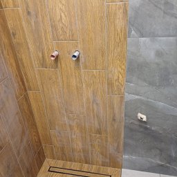 Huliailo Dmytro - Wykończona kabina prysznicowa z brązowymi płytkami imitującymi drewno na ścianach i podłodze, wbudowany odpływ liniowy, widoczne wystające rurki instalacji wodnej i fragment ściany wyłożonej...