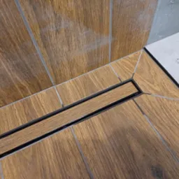 Ukośne ujęcie podłogi prysznica z odpływem liniowym, wyłożonej płytkami imitującymi drewno, z szarymi fugami i fragmentem szarej ściany w tle.