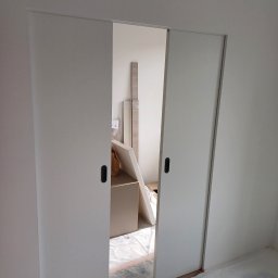 Huliailo Dmytro - Białe, przesuwne drzwi w minimalistycznym wnętrzu. Widoczne czarne uchwyty i fragmenty pomieszczenia w trakcie wykańczania, zabezpieczona podłoga folią.