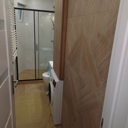 Huliailo Dmytro - Nowoczesna łazienka z kabiną prysznicową, pralką i dekoracyjną ścianą z drewnopodobnych paneli z geometrycznym wzorem. Widok zza uchylonych drzwi.