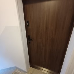 Huliailo Dmytro - Zamknięte, ciemnobrązowe drzwi wejściowe z zamkiem i klamką na tle białej ściany i fragmentu posadzki. Minimalistyczny design, proste linie, neutralne kolory.