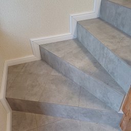 Huliailo Dmytro - Wykończone schody z szarych płytek imitujących marmur, z białymi listwami przypodłogowymi i delikatną tapetą w tle, prezentują efekt finalny prac glazurniczych.