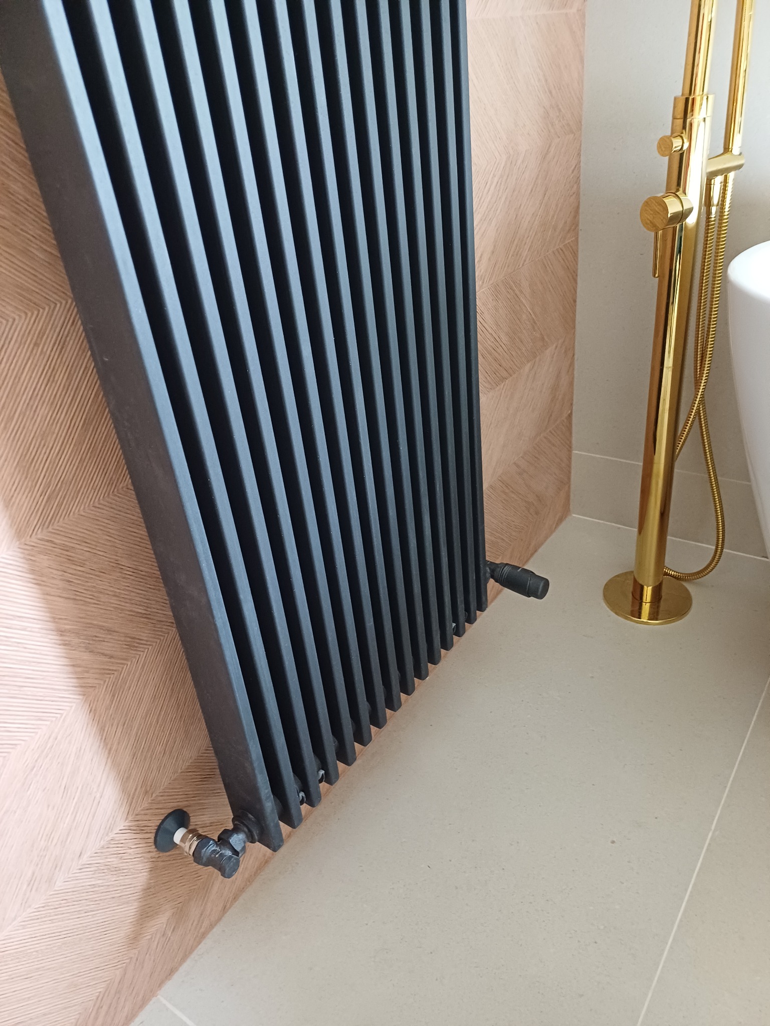 Czarny grzejnik żeberkowy na tle ściany z drewnopodobnych paneli, obok złota bateria wannowa z prysznicem na jasnej podłodze.