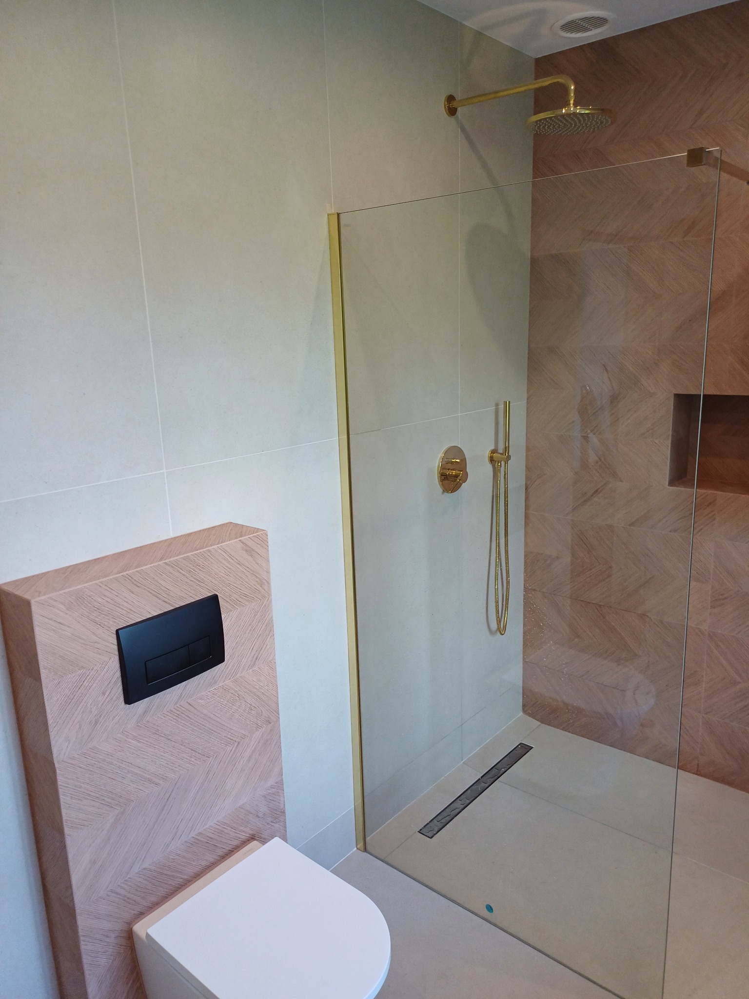 Nowoczesna łazienka z kabiną prysznicową typu walk-in, złotą armaturą i podtynkową spłuczką WC z obudową w dekorze drewna ułożonego w jodełkę.