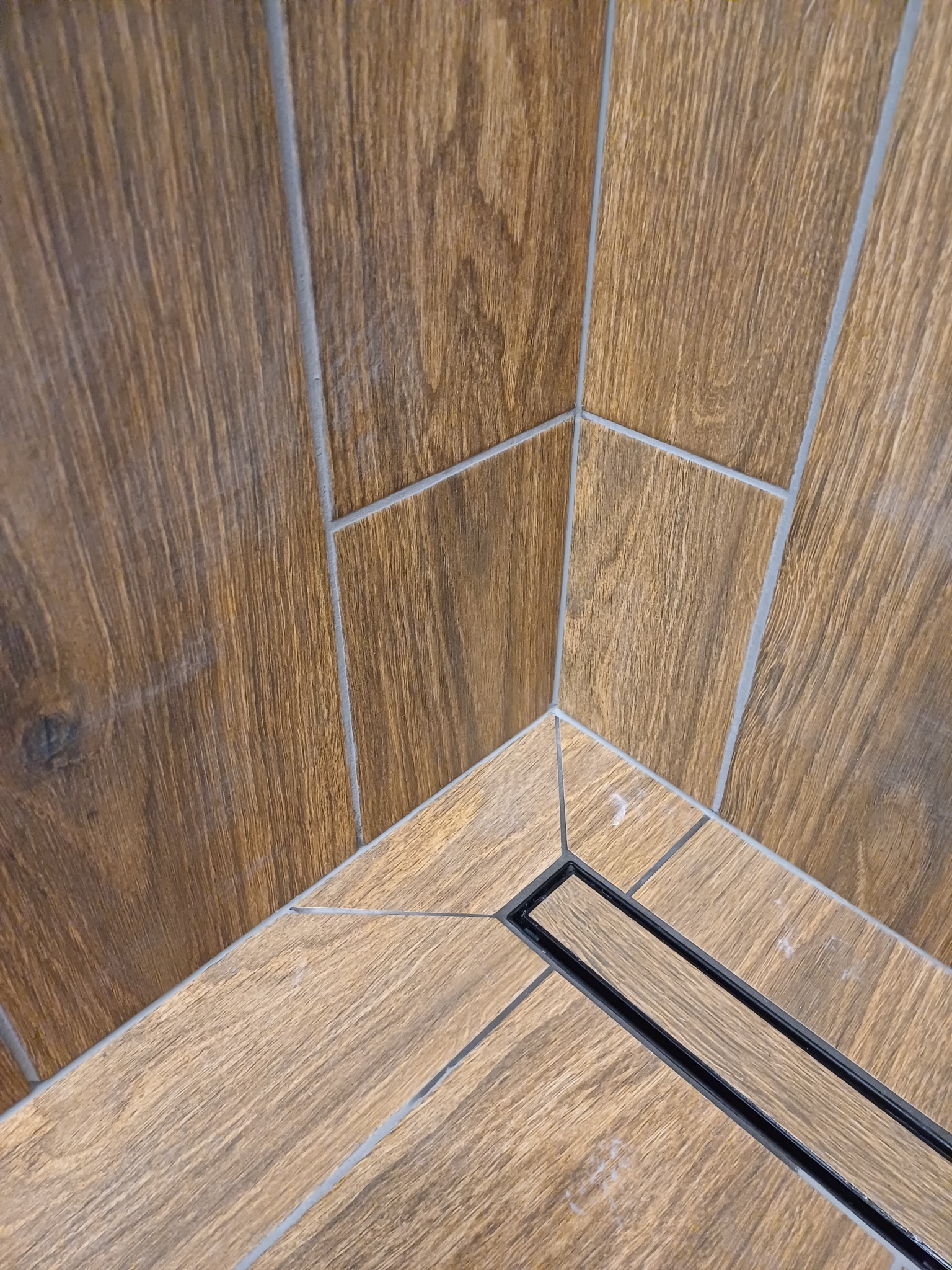 Fragment wykończonej kabiny prysznicowej z odpływem liniowym, wyłożonej płytkami imitującymi drewno w dwóch odcieniach brązu, z fugą w kolorze szarym.