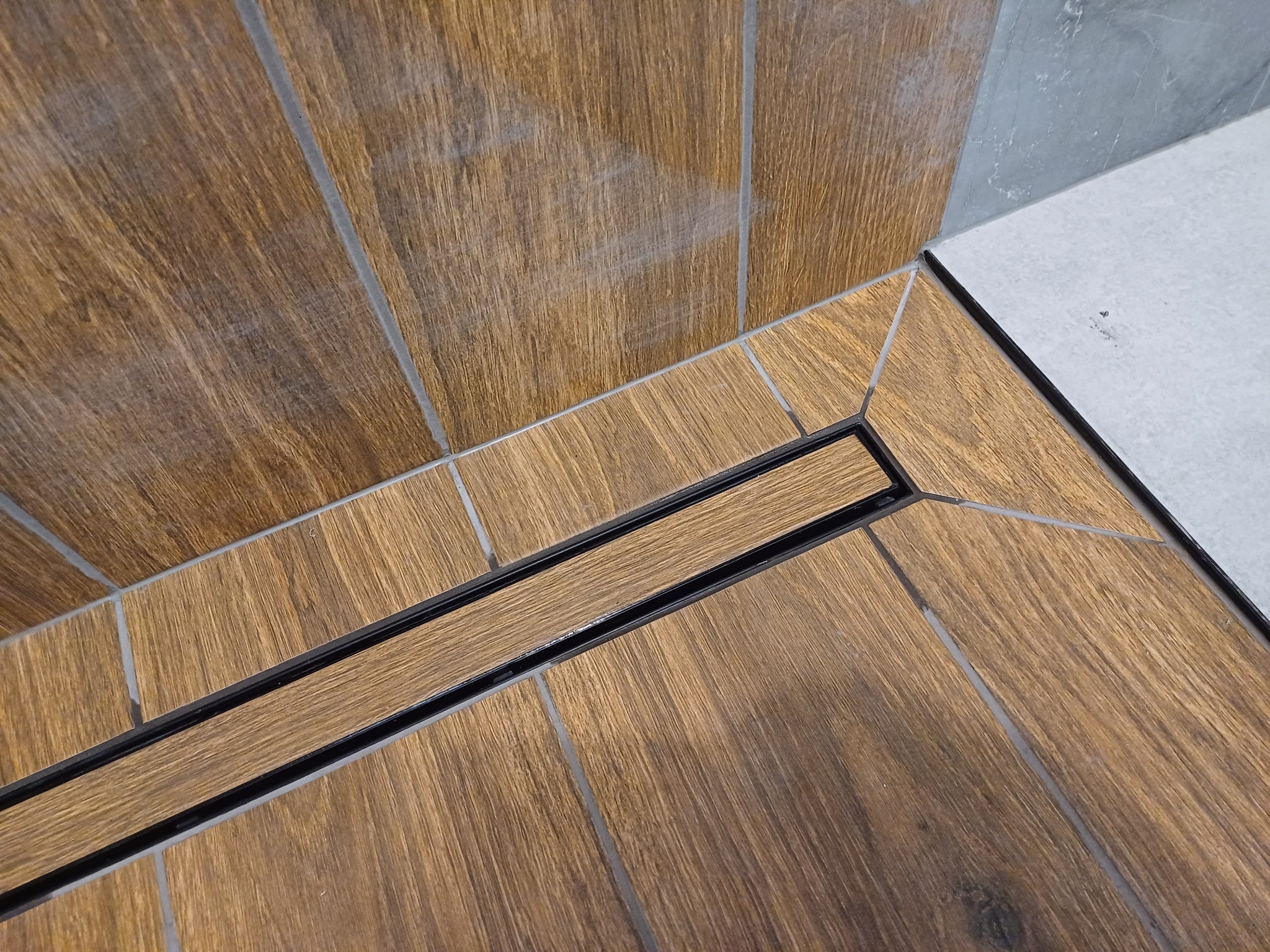 Ukośne ujęcie podłogi prysznica z odpływem liniowym, wyłożonej płytkami imitującymi drewno, z szarymi fugami i fragmentem szarej ściany w tle.