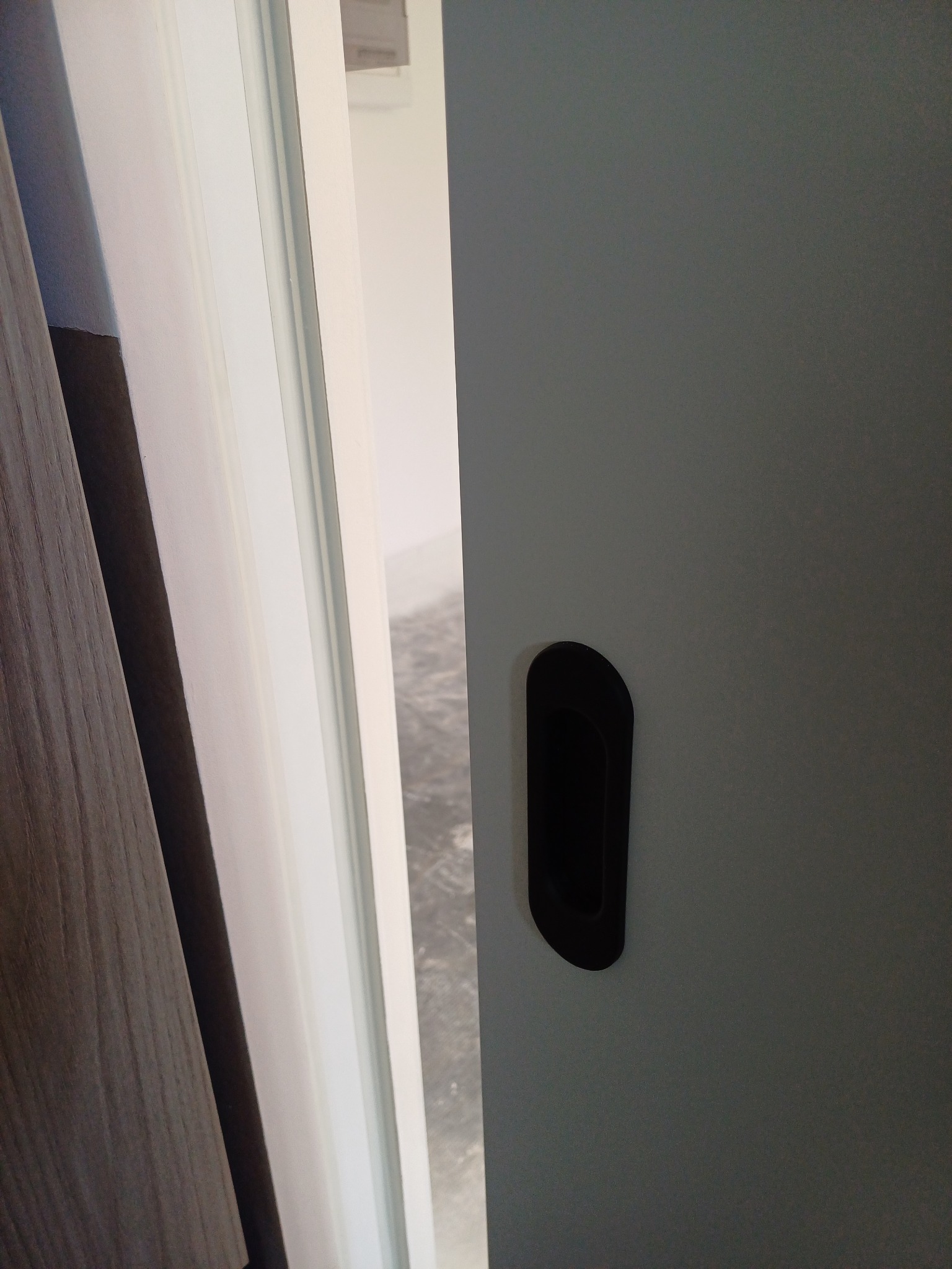 Szare drzwi przesuwne z czarnym, wpuszczanym uchwytem, widoczne z bliska. W tle fragment podłogi i ściany.