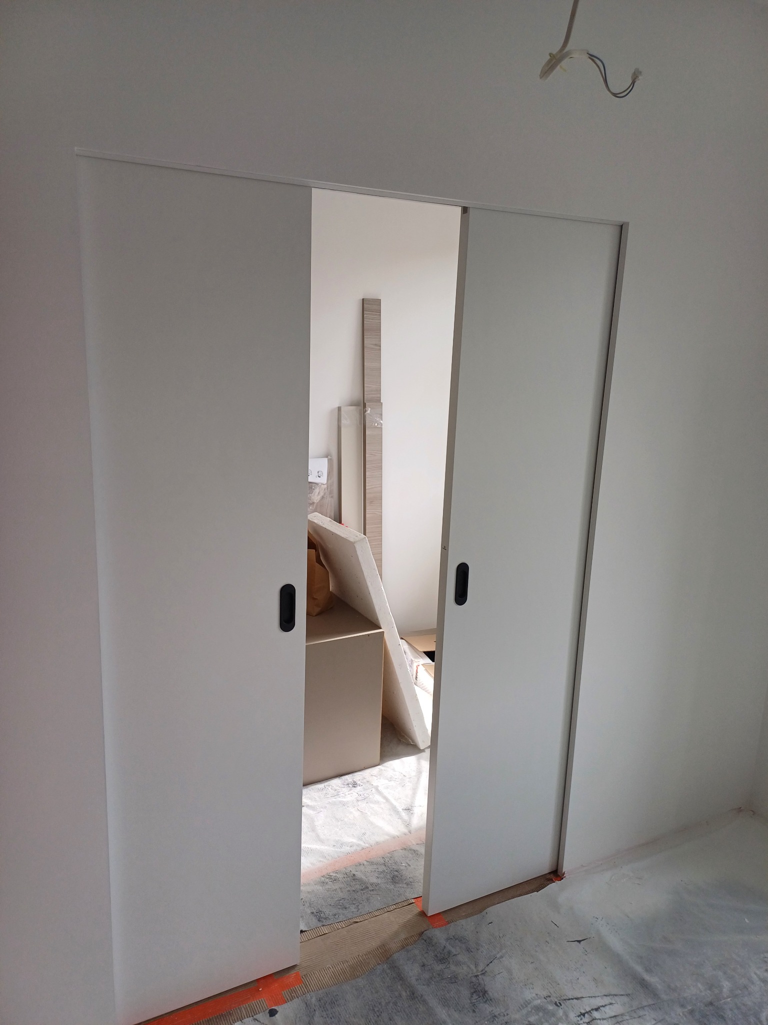 Białe, przesuwne drzwi w minimalistycznym wnętrzu. Widoczne czarne uchwyty i fragmenty pomieszczenia w trakcie wykańczania, zabezpieczona podłoga folią.