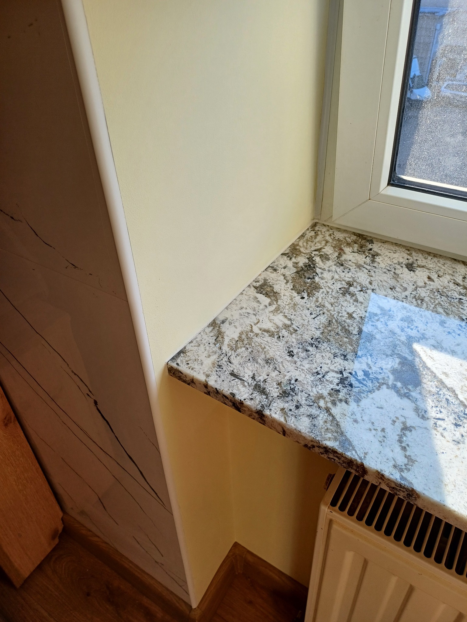 Granitowy parapet przy oknie PCV, kremowa ściana i dekoracyjna okładzina ścienna. Niski grzejnik pod parapetem. Ukończone wykończenie wnęki okiennej.