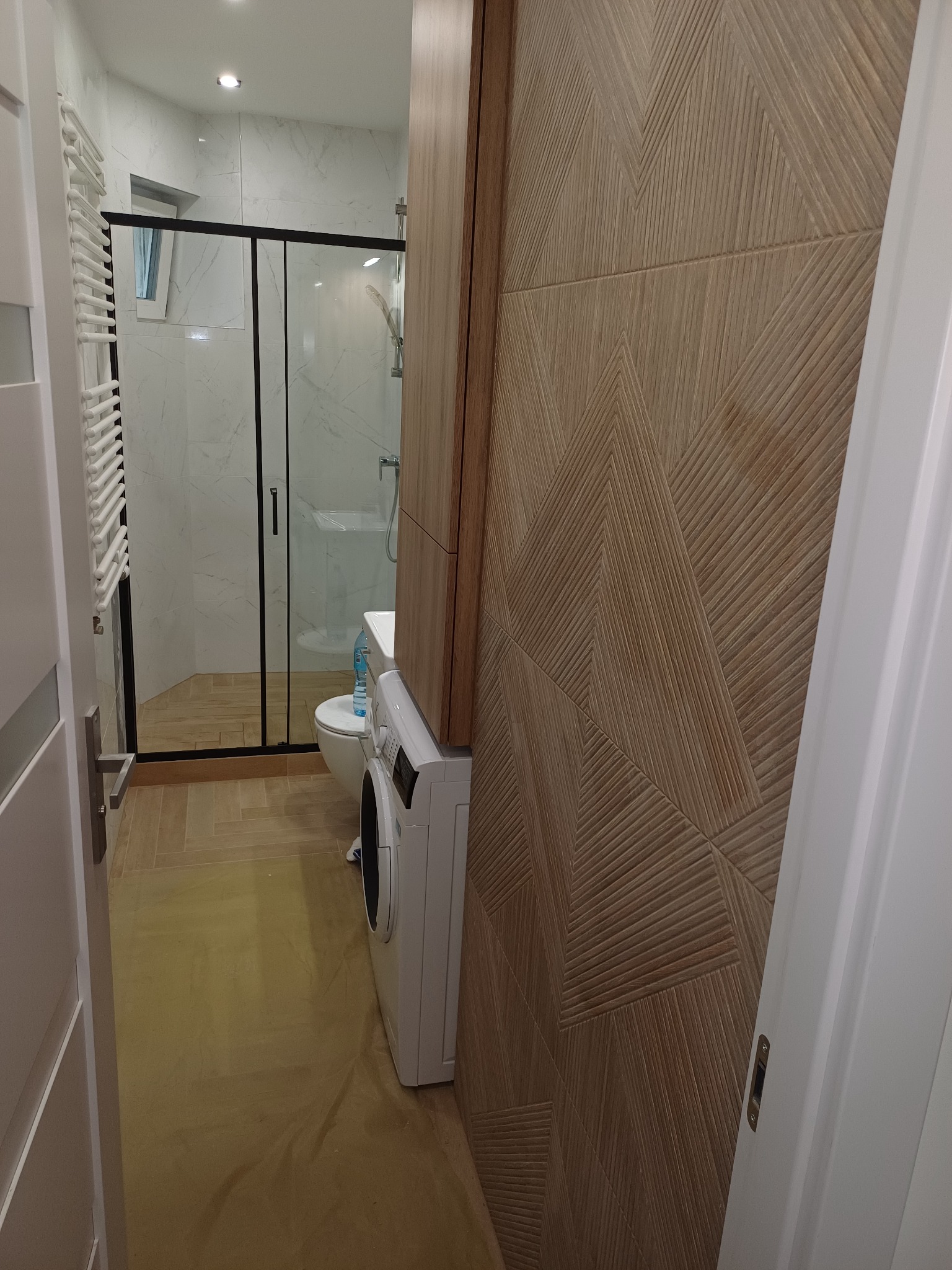 Nowoczesna łazienka z kabiną prysznicową, pralką i dekoracyjną ścianą z drewnopodobnych paneli z geometrycznym wzorem. Widok zza uchylonych drzwi.