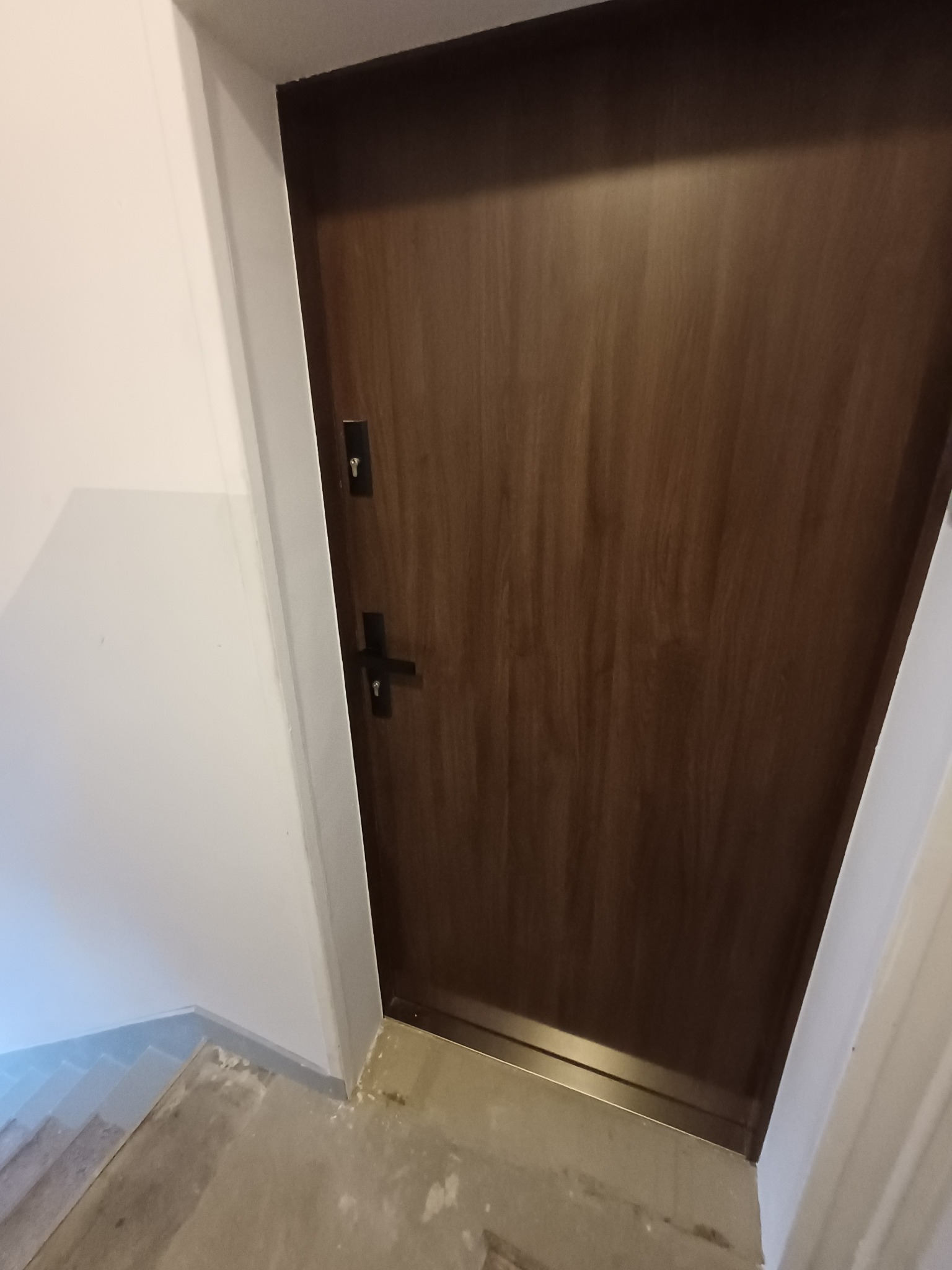 Zamknięte, ciemnobrązowe drzwi wejściowe z zamkiem i klamką na tle białej ściany i fragmentu posadzki. Minimalistyczny design, proste linie, neutralne kolory.