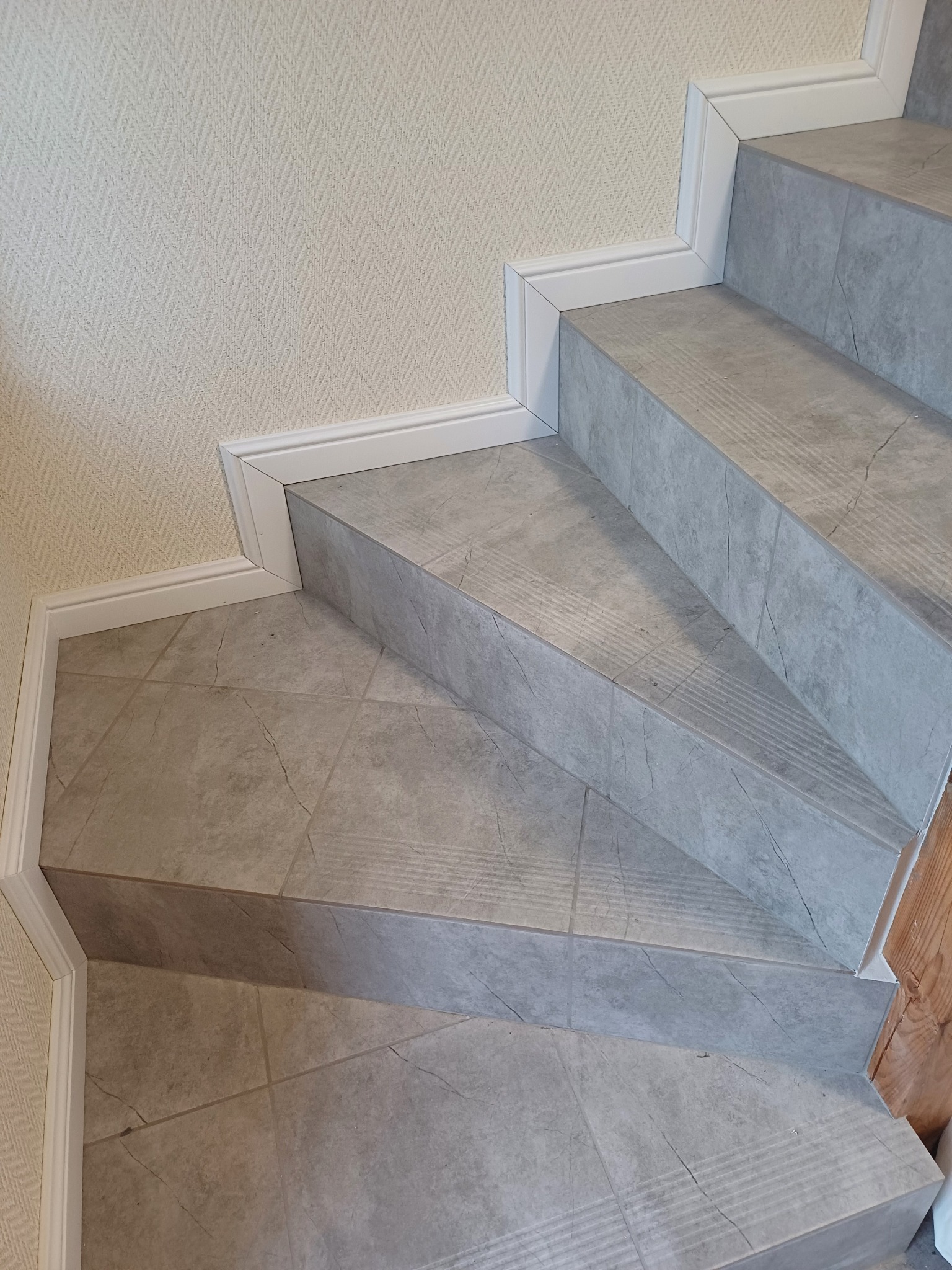 Wykończone schody z szarych płytek imitujących marmur, z białymi listwami przypodłogowymi i delikatną tapetą w tle, prezentują efekt finalny prac glazurniczych.