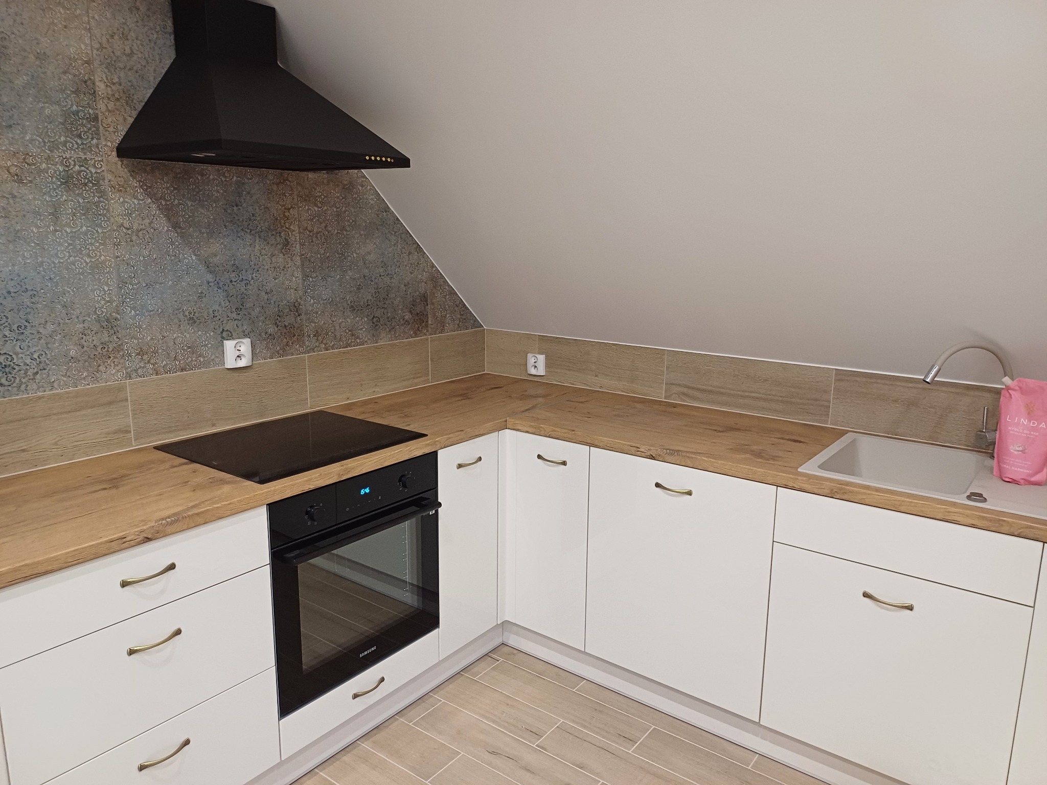 Nowoczesna kuchnia na poddaszu: białe szafki z drewnianym blatem, czarna płyta i okap. Ściana wykończona płytkami w stylu vintage.