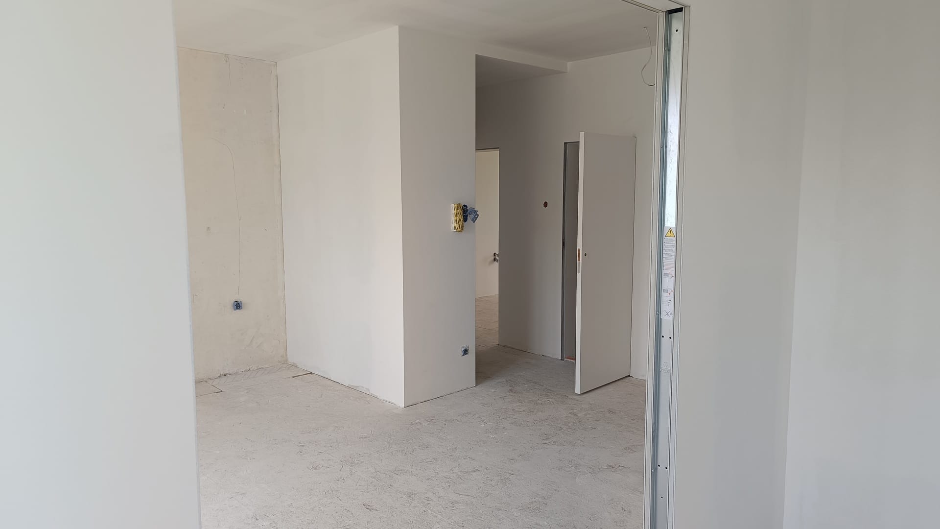 Wnętrze w trakcie prac wykończeniowych: gładkie, białe ściany, widoczne puszki elektryczne, metalowa futryna. Podłoga surowa, przygotowana do dalszych prac.