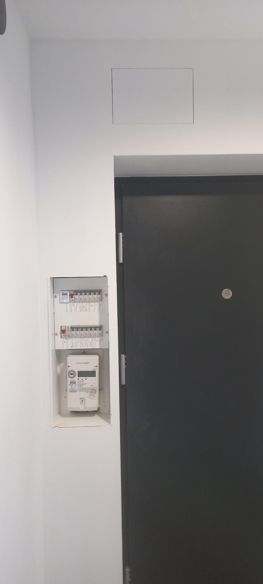 Zamontowana skrzynka elektryczna z licznikiem w białej wnęce obok ciemnych drzwi wejściowych. Widoczna instalacja elektryczna w mieszkaniu po remoncie.