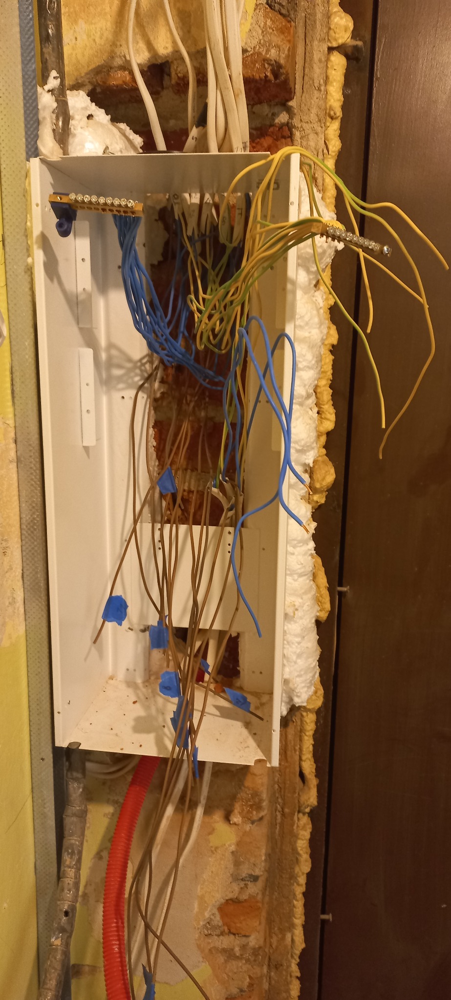 Otwarta skrzynka elektryczna z widocznymi przewodami w różnych kolorach, zamontowana na ścianie z izolacją piankową. Widoczne fragmenty instalacji elektrycznej.