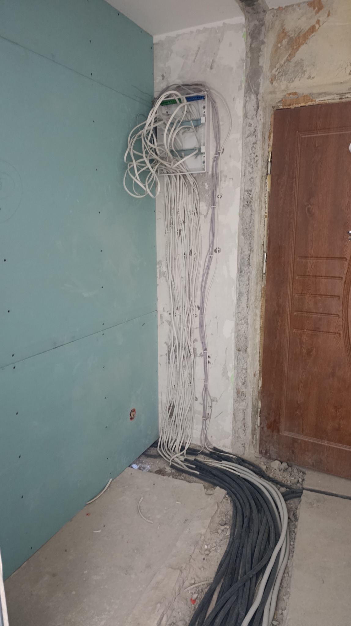 Rozdzielnica elektryczna z licznymi przewodami na ścianie obok płyty karton-gips, widoczne kable na podłodze, surowy stan wykończenia wnętrza.