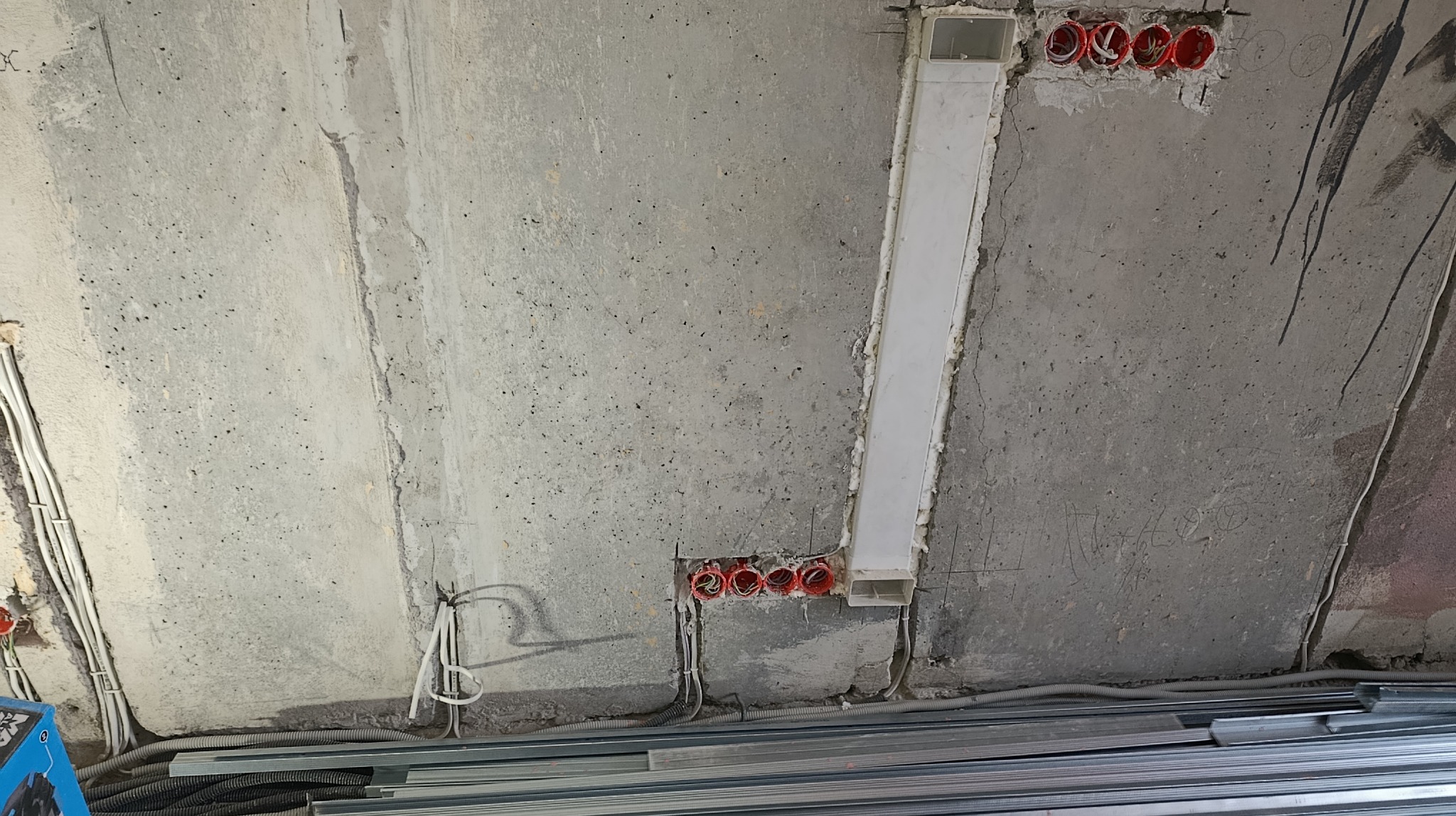 Betonowy sufit w trakcie prac instalacyjnych: widoczne kable, puszki elektryczne w kolorze czerwonym, kanał wentylacyjny i profile metalowe na podłodze.