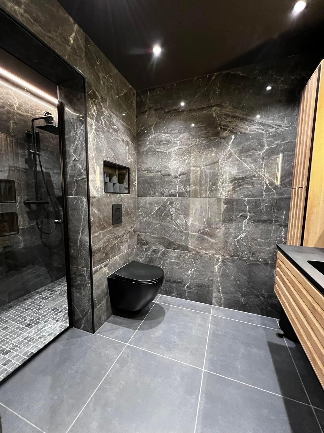 Nowoczesna łazienka w odcieniach szarości z czarną armaturą: wisząca toaleta, kabina prysznicowa z deszczownicą i umywalka z szafką z drewnianymi lamelami. Ściany wyłożone płytkami imitującymi...