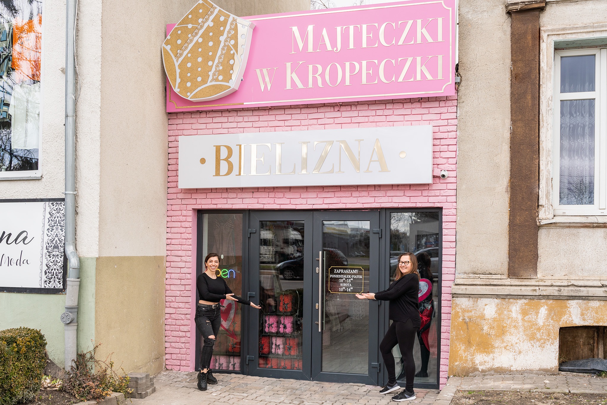 Fasada sklepu z bielizną 'Majteczki w Kropeczki' z różową cegłą i szyldami, dwie kobiety w czarnych ubraniach zapraszają do środka, prezentując godziny otwarcia na drzwiach.