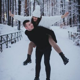 Fotografia Oskar Borzestowski - Uśmiechnięta para w zimowej scenerii: mężczyzna niesie kobietę na barana w zasypanym śniegiem lesie, ona rozkłada ręce jak skrzydła, oboje w ciepłych, zimowych ubraniach i butach.
