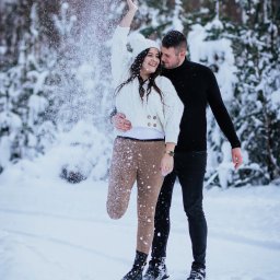 Fotografia Oskar Borzestowski - Uśmiechnięta para w zimowej scenerii: kobieta rzuca śnieg w górę, mężczyzna obejmuje ją czule, oboje ubrani w ciepłe, kontrastujące stroje na tle ośnieżonych drzew.