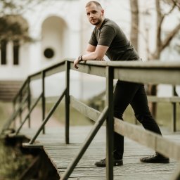 Fotografia Oskar Borzestowski - Mężczyzna oparty o drewnianą barierkę mostu, w tle budynek z łukowym wejściem, stonowana kolorystyka.