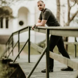 Mężczyzna oparty o drewnianą barierkę mostu, w tle budynek z łukowym wejściem, stonowana kolorystyka.