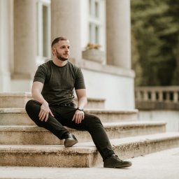 Fotografia Oskar Borzestowski - Mężczyzna w czarnym ubraniu siedzi na kamiennych schodach klasycznego budynku z kolumnami, patrząc w dal.