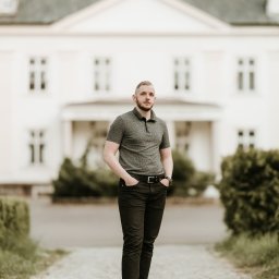 Fotografia Oskar Borzestowski - Mężczyzna w szarej koszulce polo i czarnych spodniach pozuje na brukowanej ścieżce przed białym budynkiem z szarym dachem.