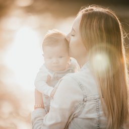 Fotografia Oskar Borzestowski - Matka całuje w czoło trzymane na rękach niemowlę ubrane w jasne ubranko, ciepłe światło zachodzącego słońca tworzy miękkie tło.