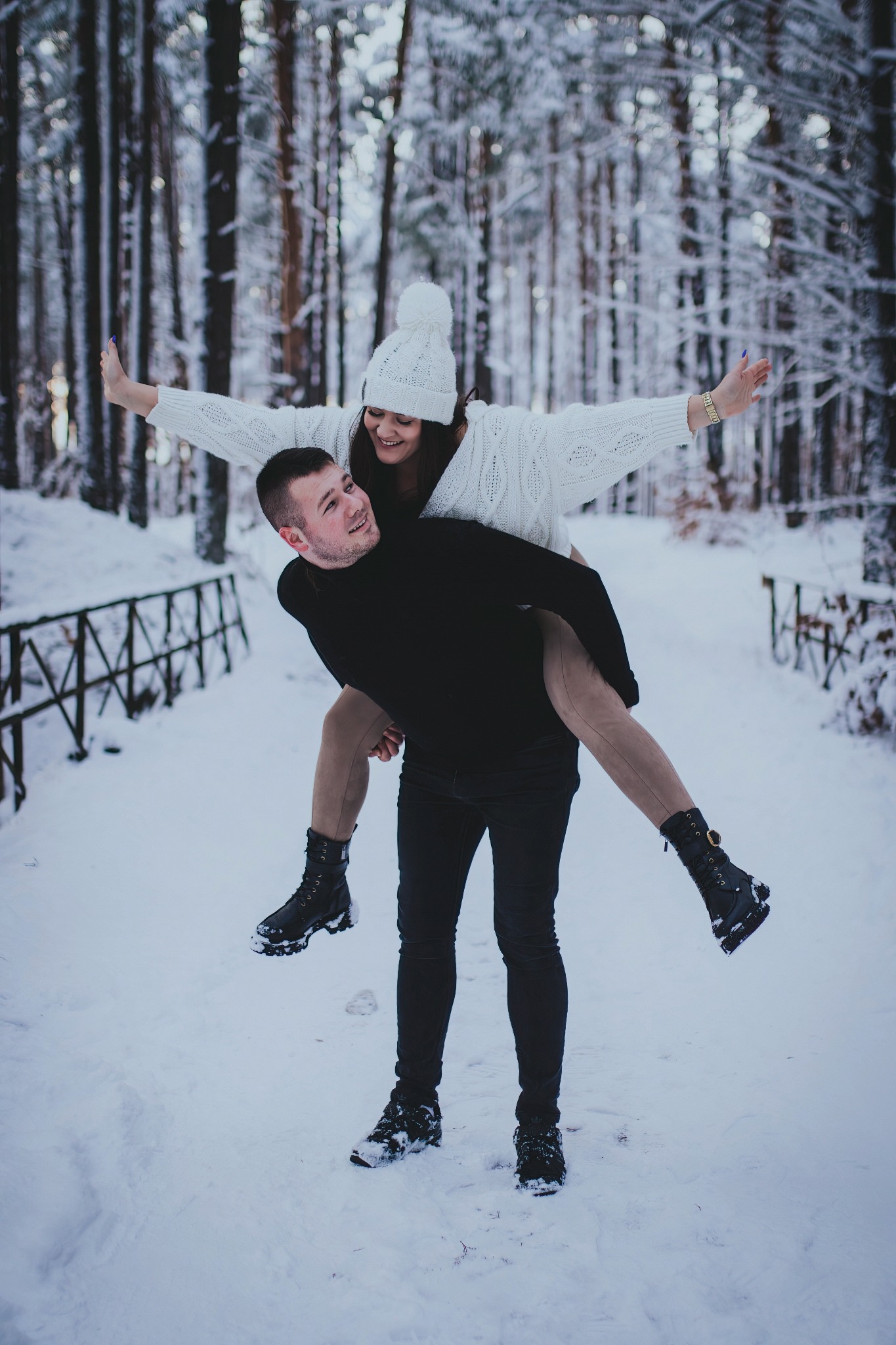 Uśmiechnięta para w zimowej scenerii: mężczyzna niesie kobietę na barana w zasypanym śniegiem lesie, ona rozkłada ręce jak skrzydła, oboje w ciepłych, zimowych ubraniach i butach.