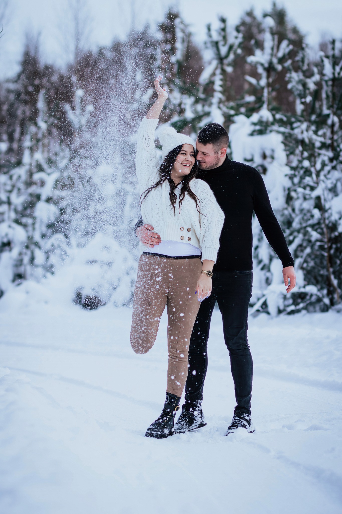 Uśmiechnięta para w zimowej scenerii: kobieta rzuca śnieg w górę, mężczyzna obejmuje ją czule, oboje ubrani w ciepłe, kontrastujące stroje na tle ośnieżonych drzew.