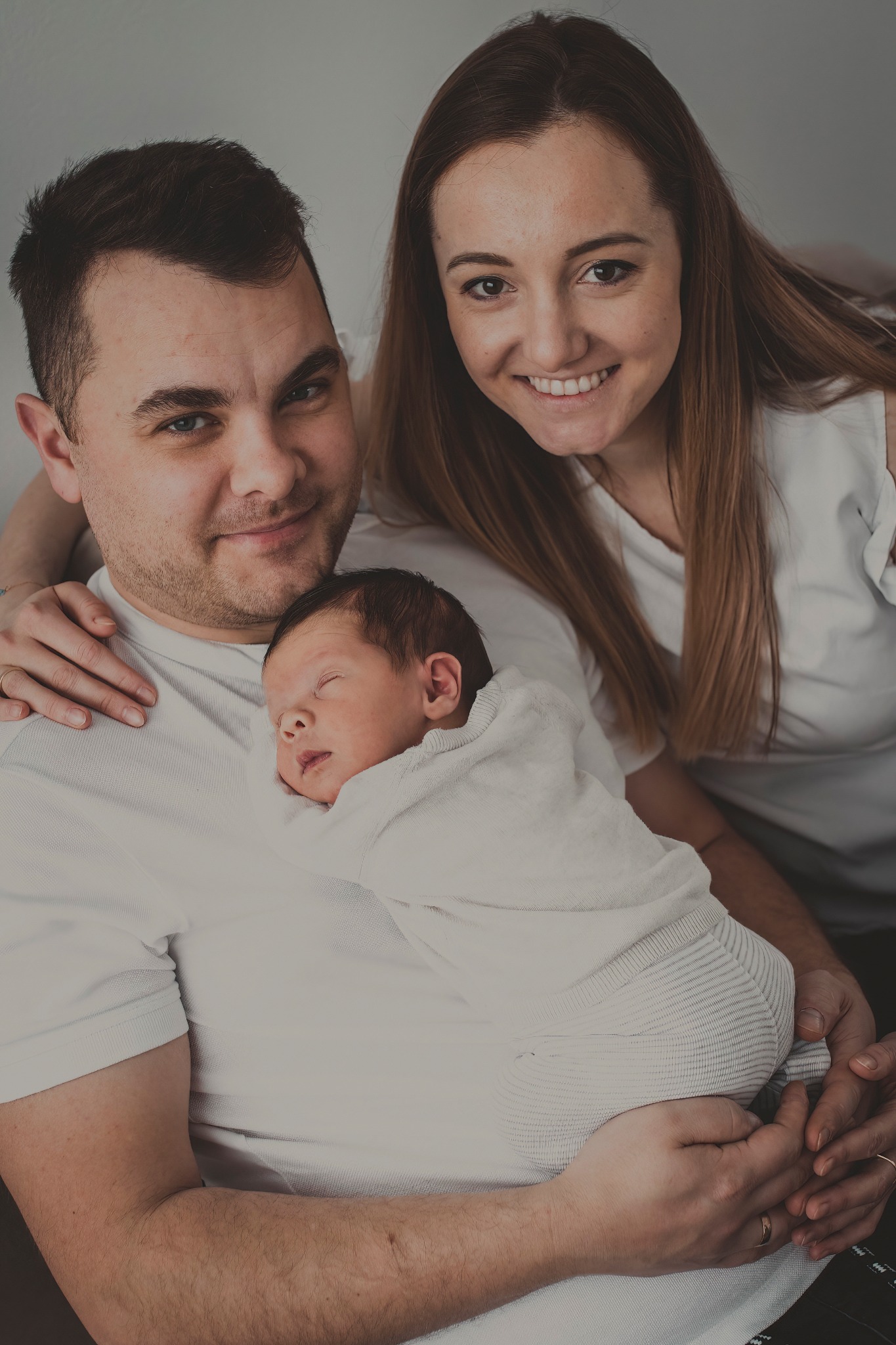 Szczęśliwa rodzina: tata trzyma noworodka zawiniętego w biały kocyk, mama obejmuje ich oboje, uśmiechając się do kamery.