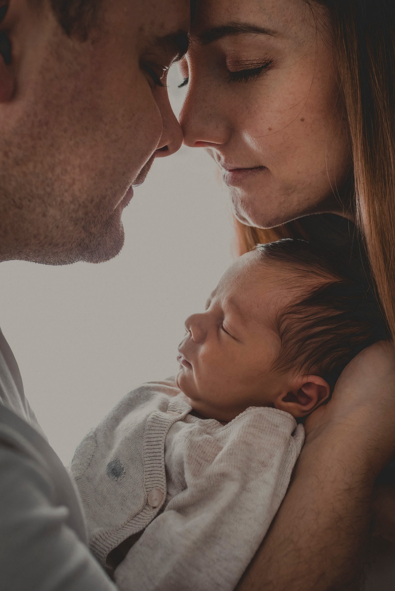 Rodzice dotykają się nosami nad śpiącym noworodkiem ubranym w kremowy sweterek, profil twarzy dziecka i zamyślone spojrzenia rodziców.