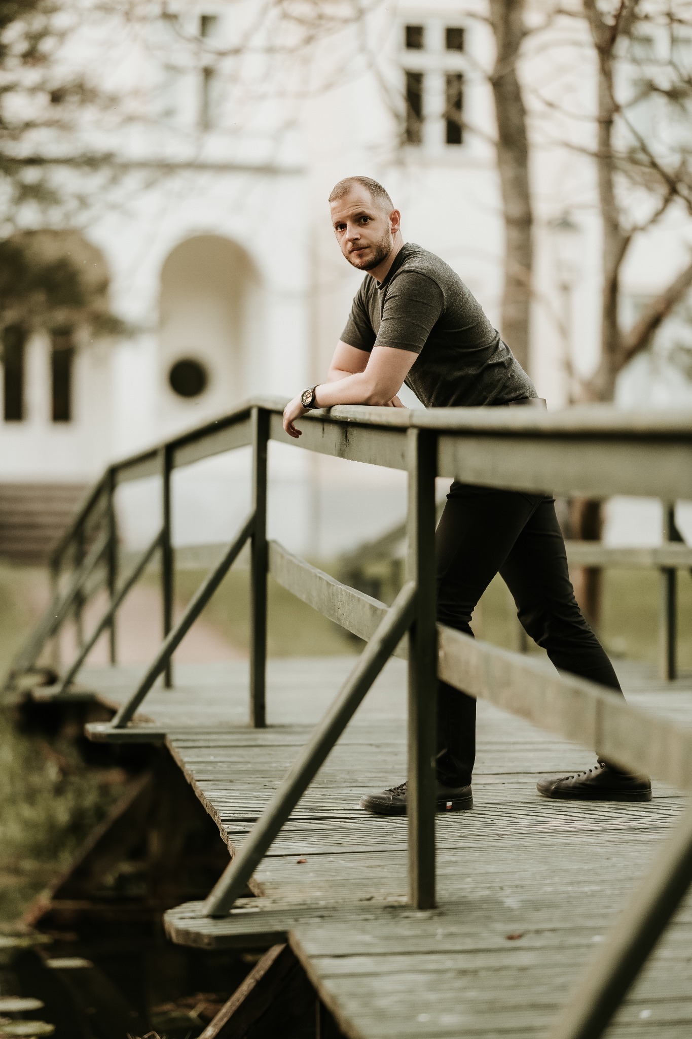 Mężczyzna oparty o drewnianą barierkę mostu, w tle budynek z łukowym wejściem, stonowana kolorystyka.